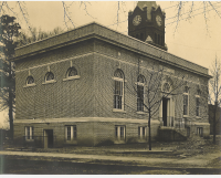 Historic sepia-toned photograph of Greenwood County Carnegie Library building, a two-story brick building with tall arched windows, a small staircase leading to a side entrance, and bare winter trees in the foreground. A large clock tower rises behind the building, partially visible above the roofline. The scene appears quiet, with an empty sidewalk and leaf-strewn ground.