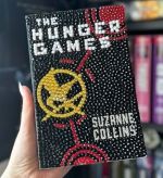 A hand holds a copy of The Hunger Games by Suzanne Collins with the entire book cover decorated in rhinestones. The rhinestones recreate the black background, the yellow mockingjay emblem, and red circular accents, giving the cover a sparkling, textured look.