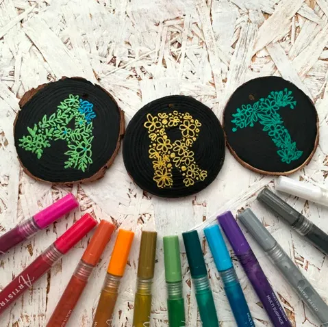 Three painted wood slices spelling out the word ‘ART’ with floral designs in green, yellow, and teal on a black background, surrounded by colorful paint markers arranged in a semicircle below.