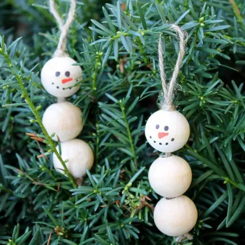 “Two handmade snowman ornaments made from stacked wooden balls with drawn-on faces and twine loops for hanging, displayed on green evergreen branches