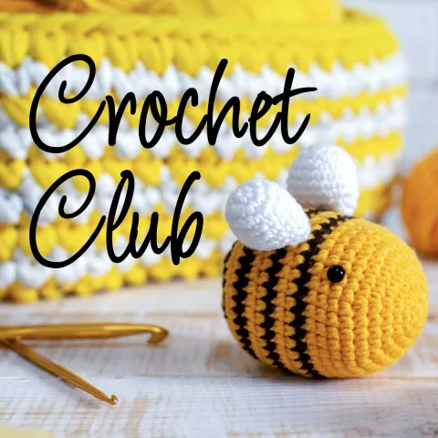 A cute crocheted yellow and black bee with white wings sits next to a gold crochet hook on a wooden surface. In the background, there’s a yellow and white crocheted basket. The words ‘Crochet Club’ are written in a flowing black script across the image.